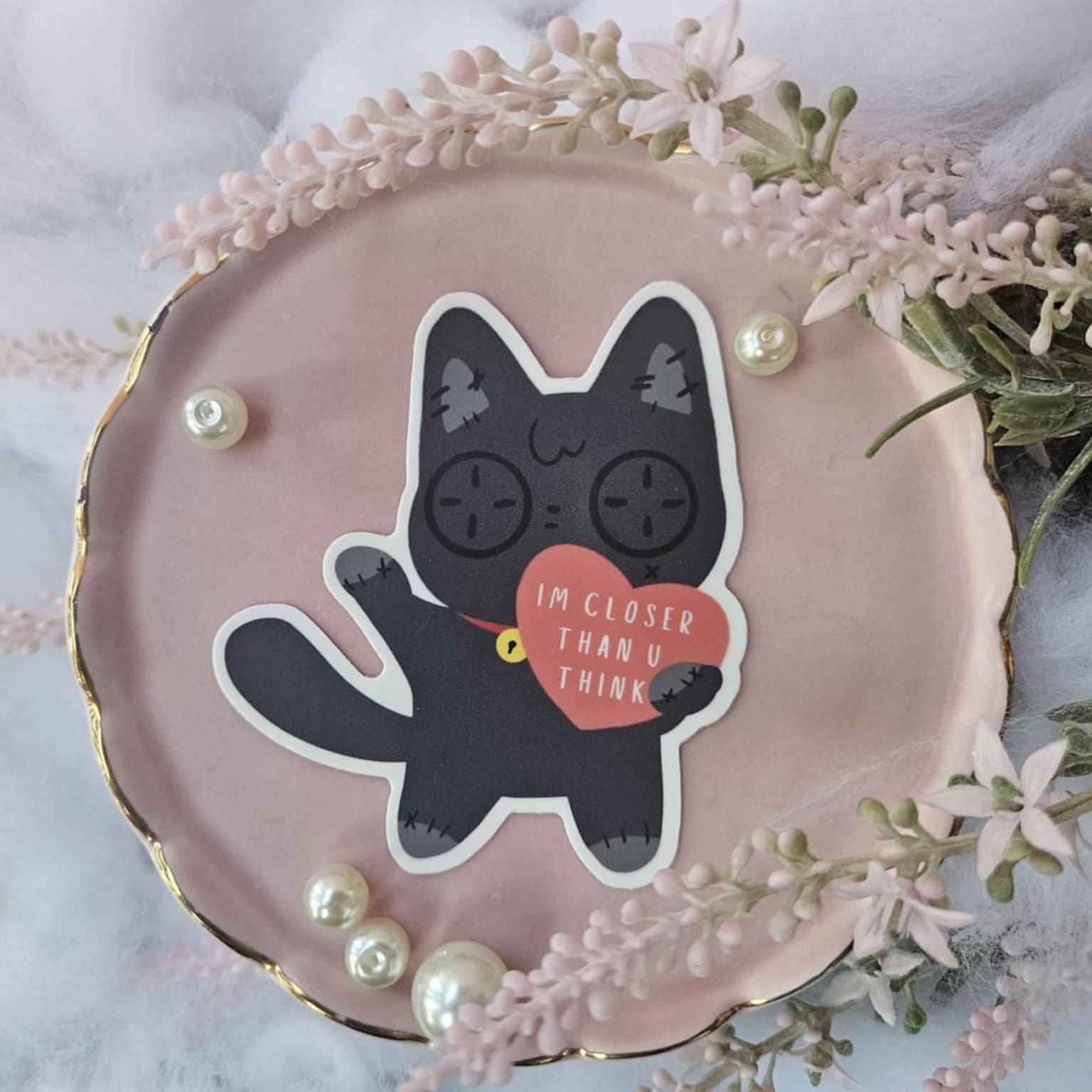 Pink decorative plate with a button eyed black cat sticker illustration and text, surrounded by flowers and pearls.