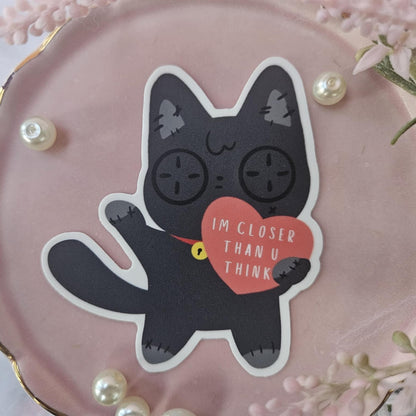 Button eye black cat sticker with a heart with text on it "I'm closer than u think" on a pink plate background