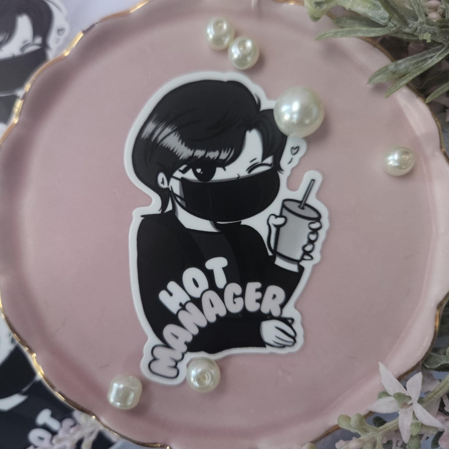 Pink round object with a black and white illustration of a person wearing a mask and 'HOT MANAGER' text, surrounded by pearls.