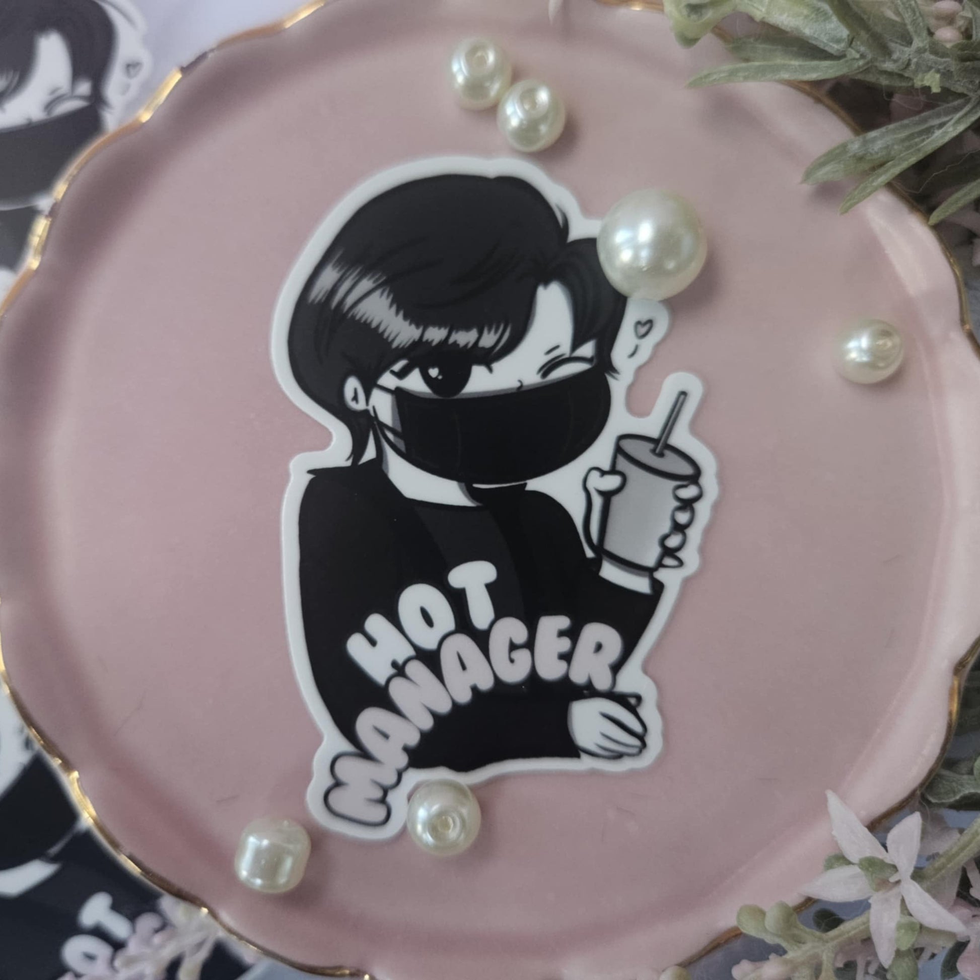 Pink round object with a black and white illustration of a person wearing a mask and 'HOT MANAGER' text, surrounded by pearls.