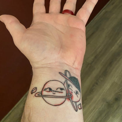 Photo of a wrist tattoo depicting: Two planet drawings next to each other; one with a black baseball hat; each with a coffee cup floating next to them.