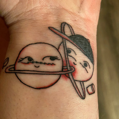 Photo of a wrist tattoo depicting close up: Two planet drawings next to each other; one with a black baseball hat; each with a coffee cup floating next to them.